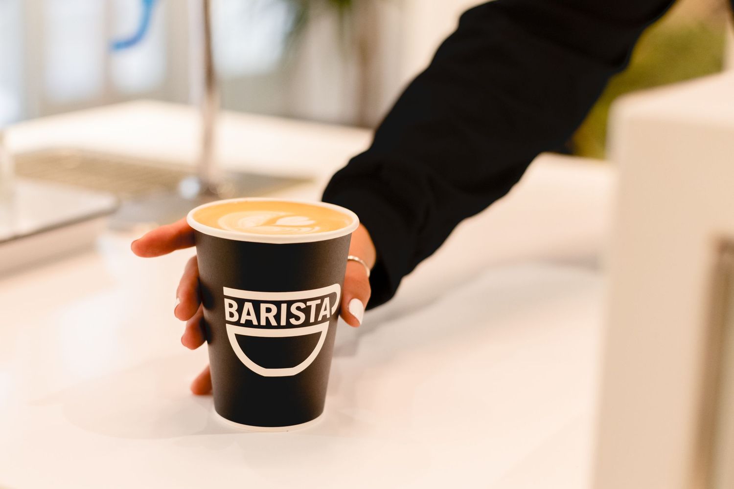 Hand in black glove holding a paper coffee cup with Barista logo against blurred light background.