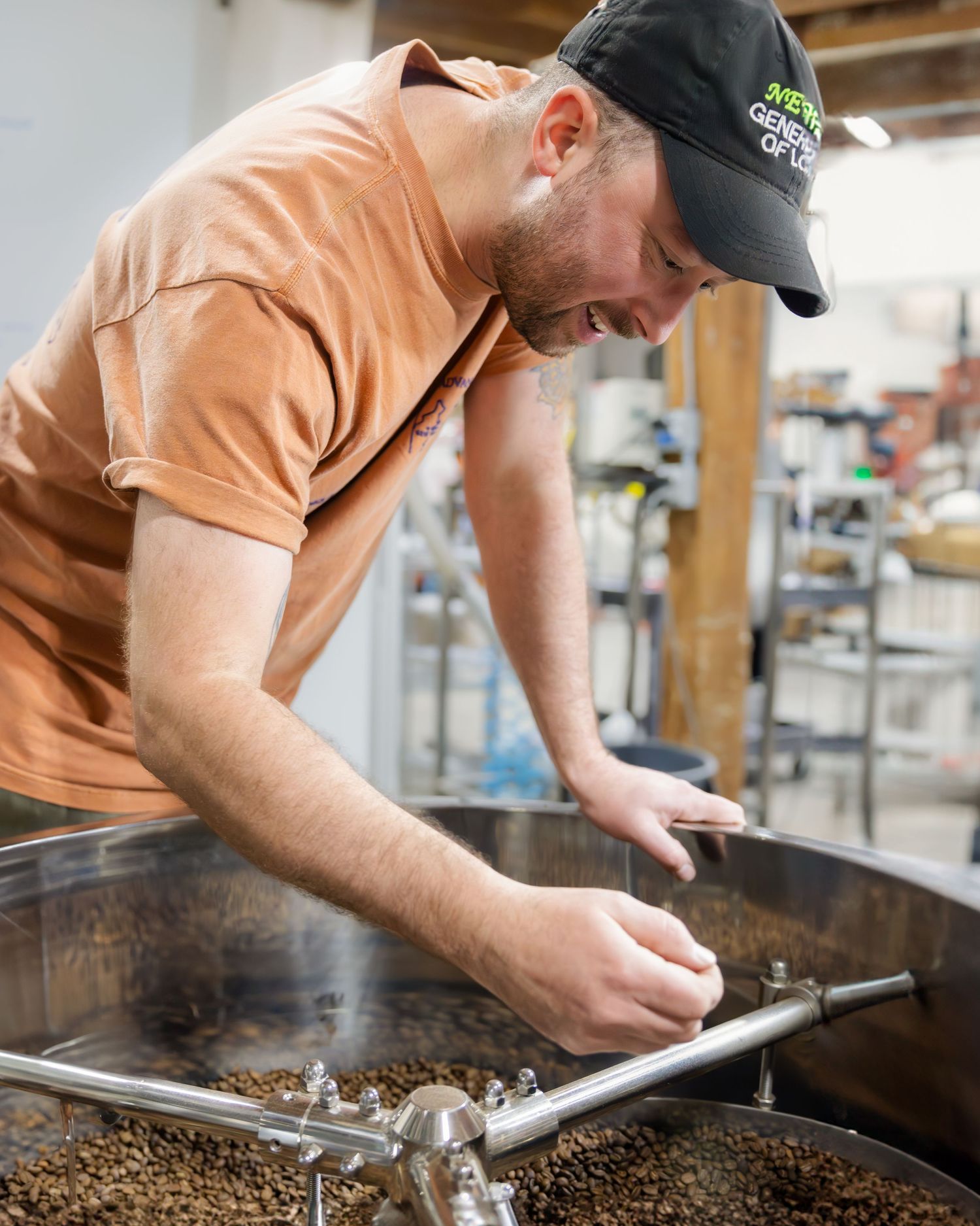 A person in a baseball cap inspects freshly roasted coffee beans in a large commercial roasting drum.