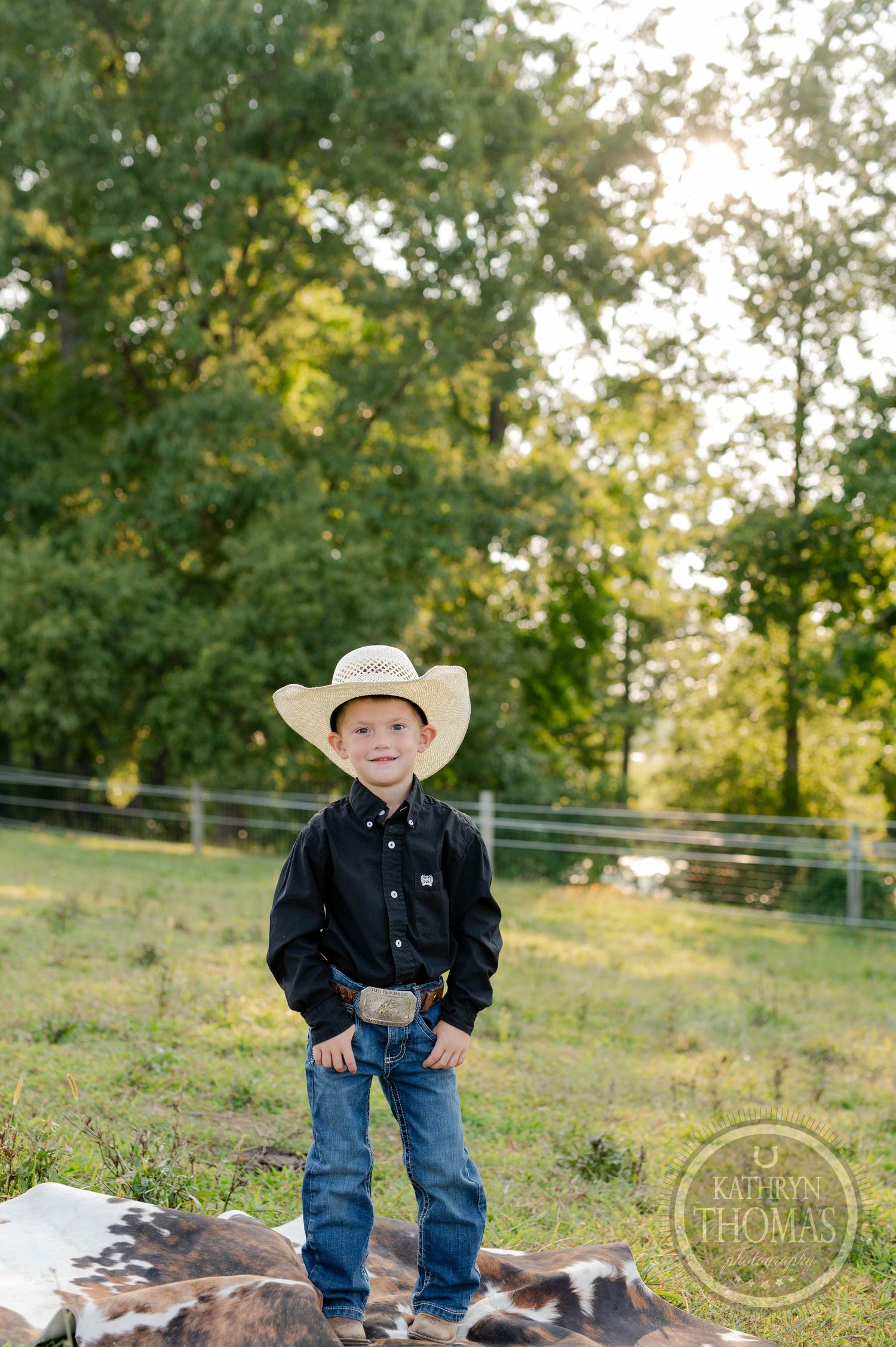 Young cowboy in hat and jeans stands confidently on spotted cowhide in rural setting.