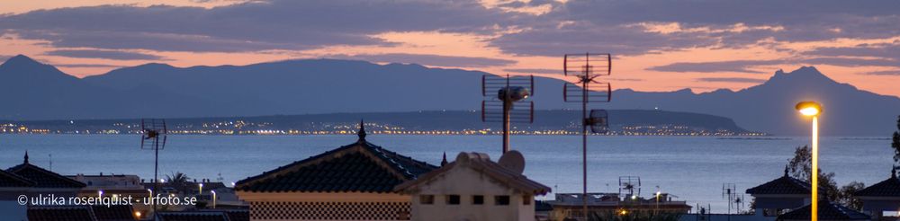 Panoramic sunset view over city rooftops with silhouetted buildings and glowing street lights.