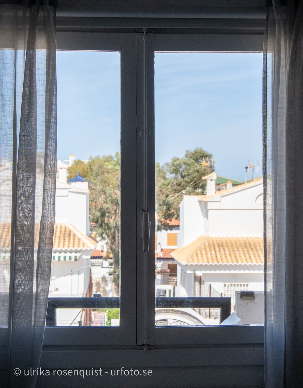 View through curtained window showing white buildings with terracotta roofs under blue sky.