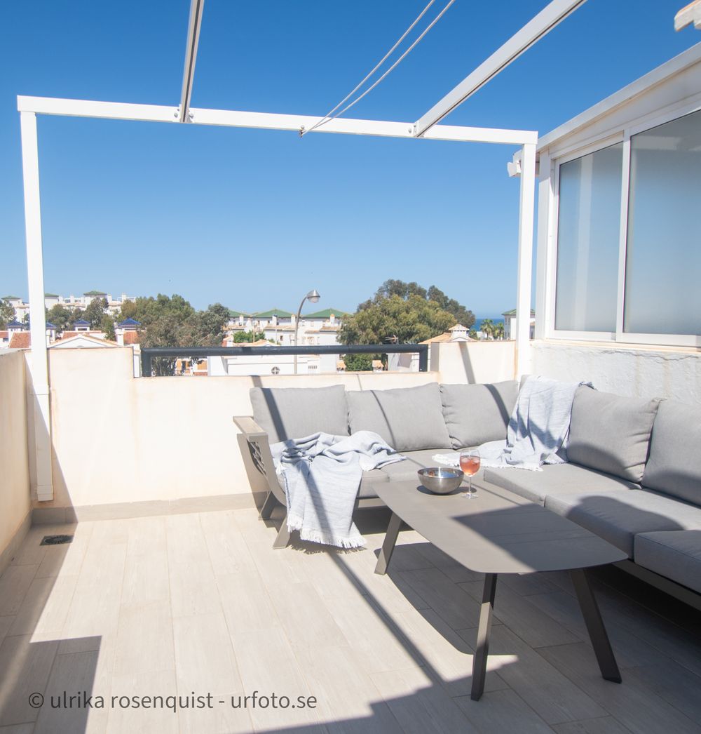 Sunny rooftop terrace with grey outdoor furniture and white pergola overlooking coastal views.
