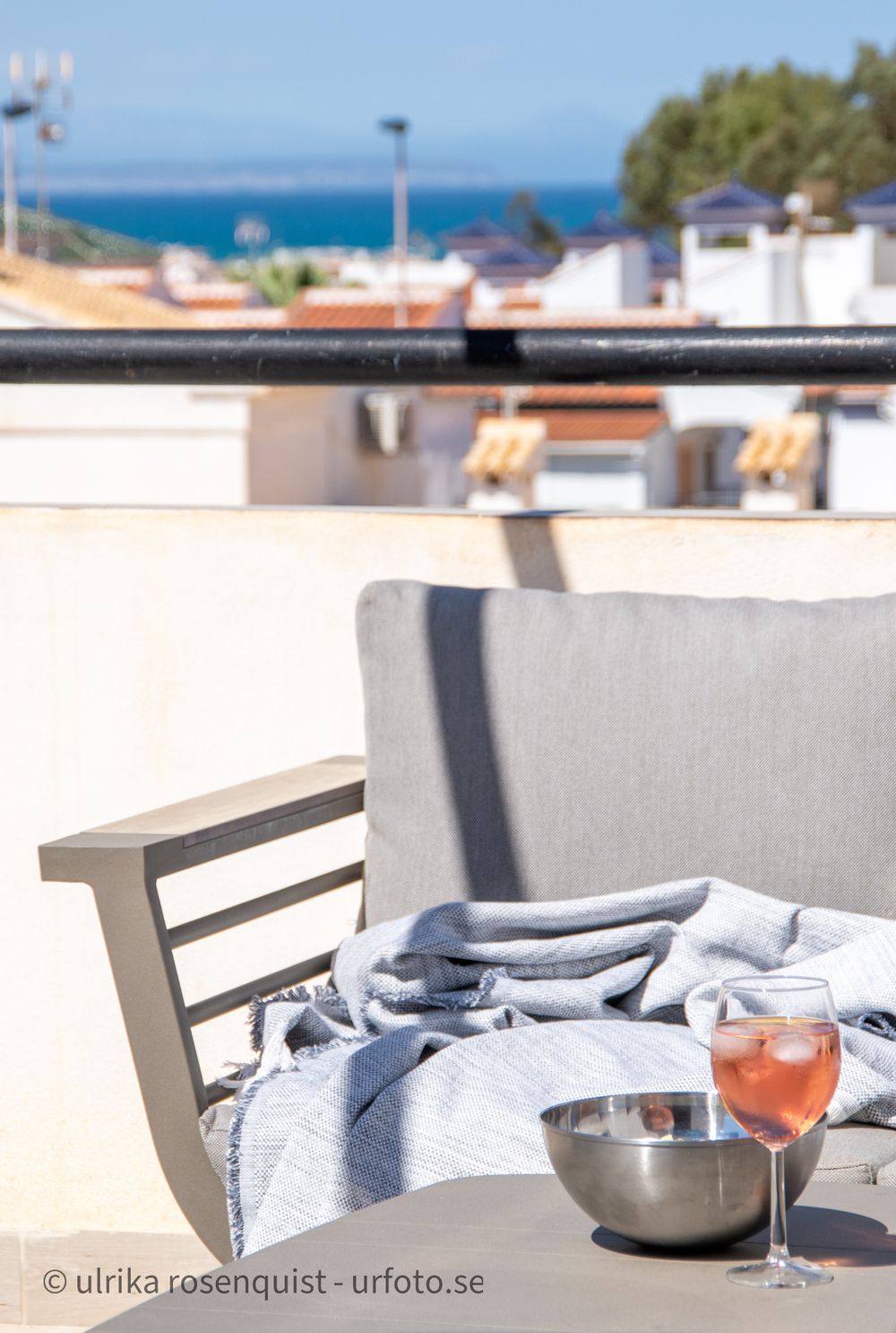 Outdoor balcony seating with grey cushions, blanket and wine glass overlooking ocean view.