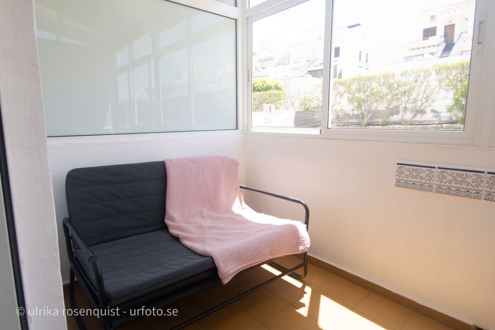 Sunlit enclosed balcony with black and pink bench seating against white walls and large windows.