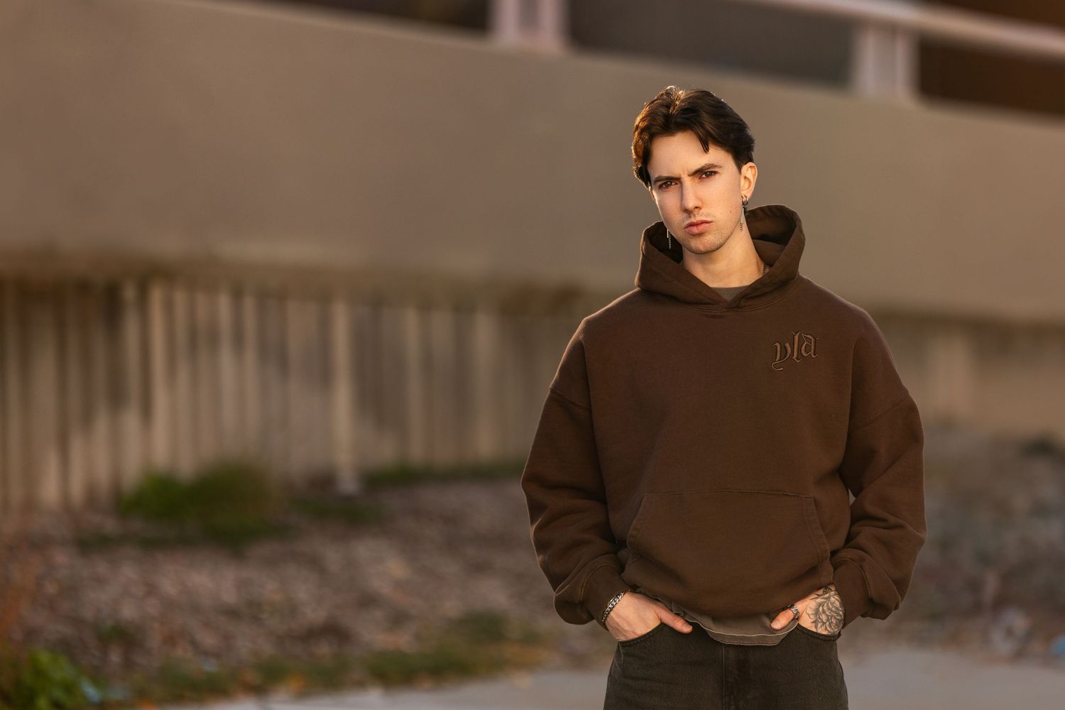 Person in brown hoodie standing outdoors during golden hour with hands in pockets.