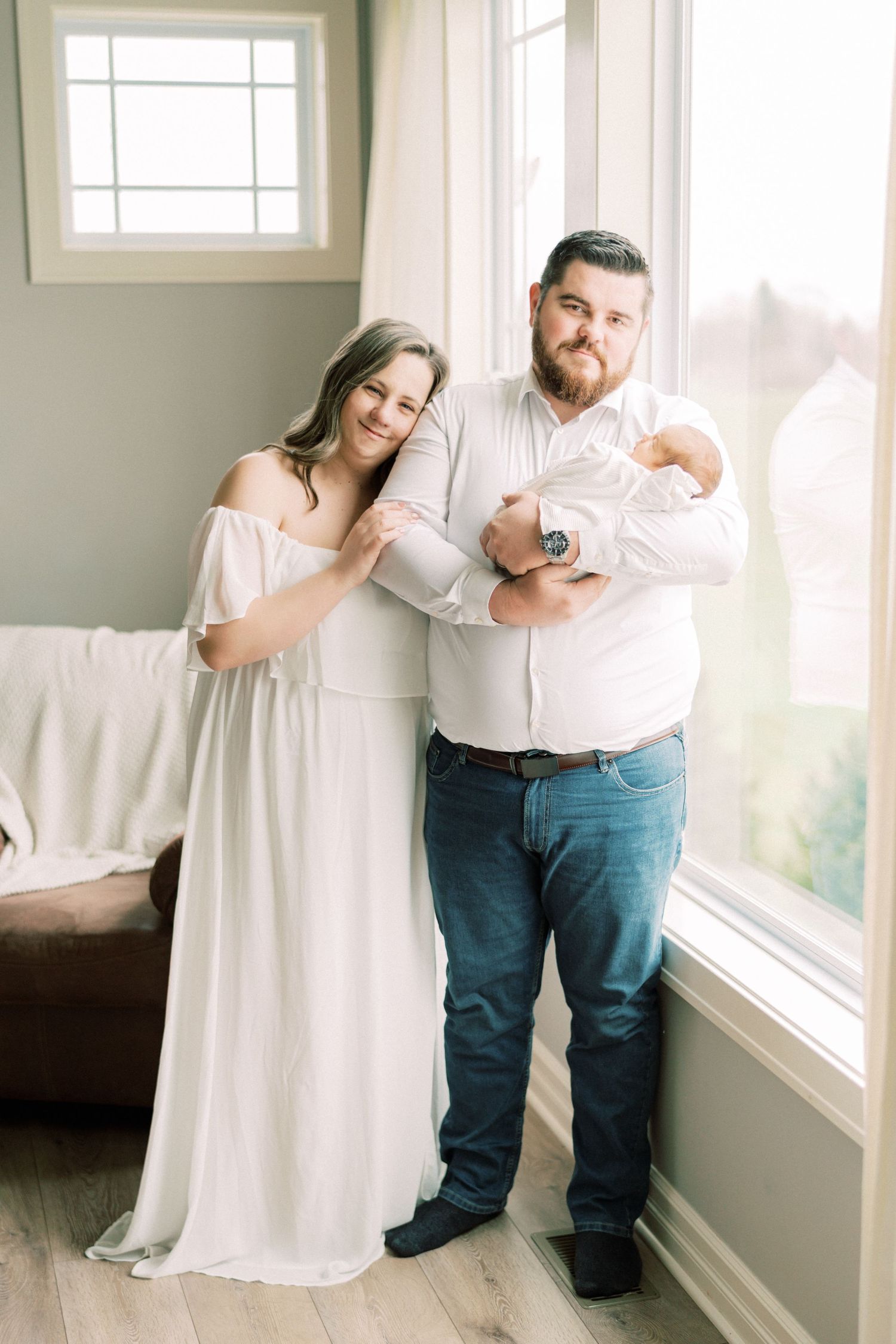 A family relaxes together on a bright white couch by large windows in a tender moment at home.