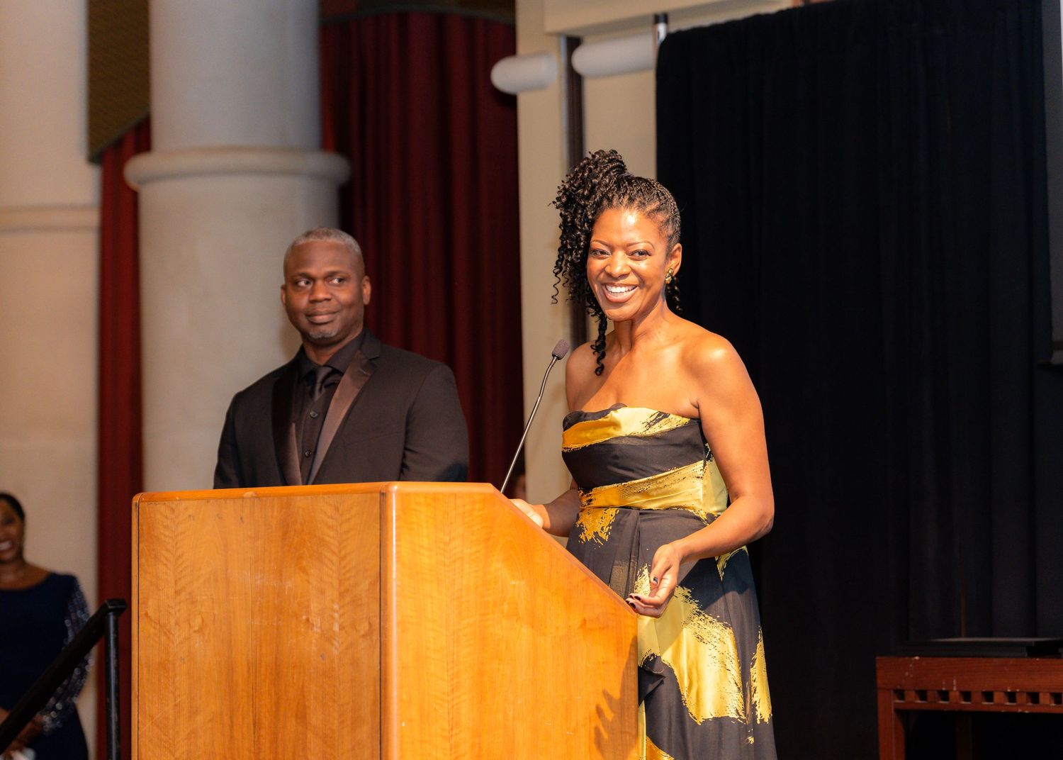 Speaker at a podium wearing an elegant yellow and black dress during a formal event.