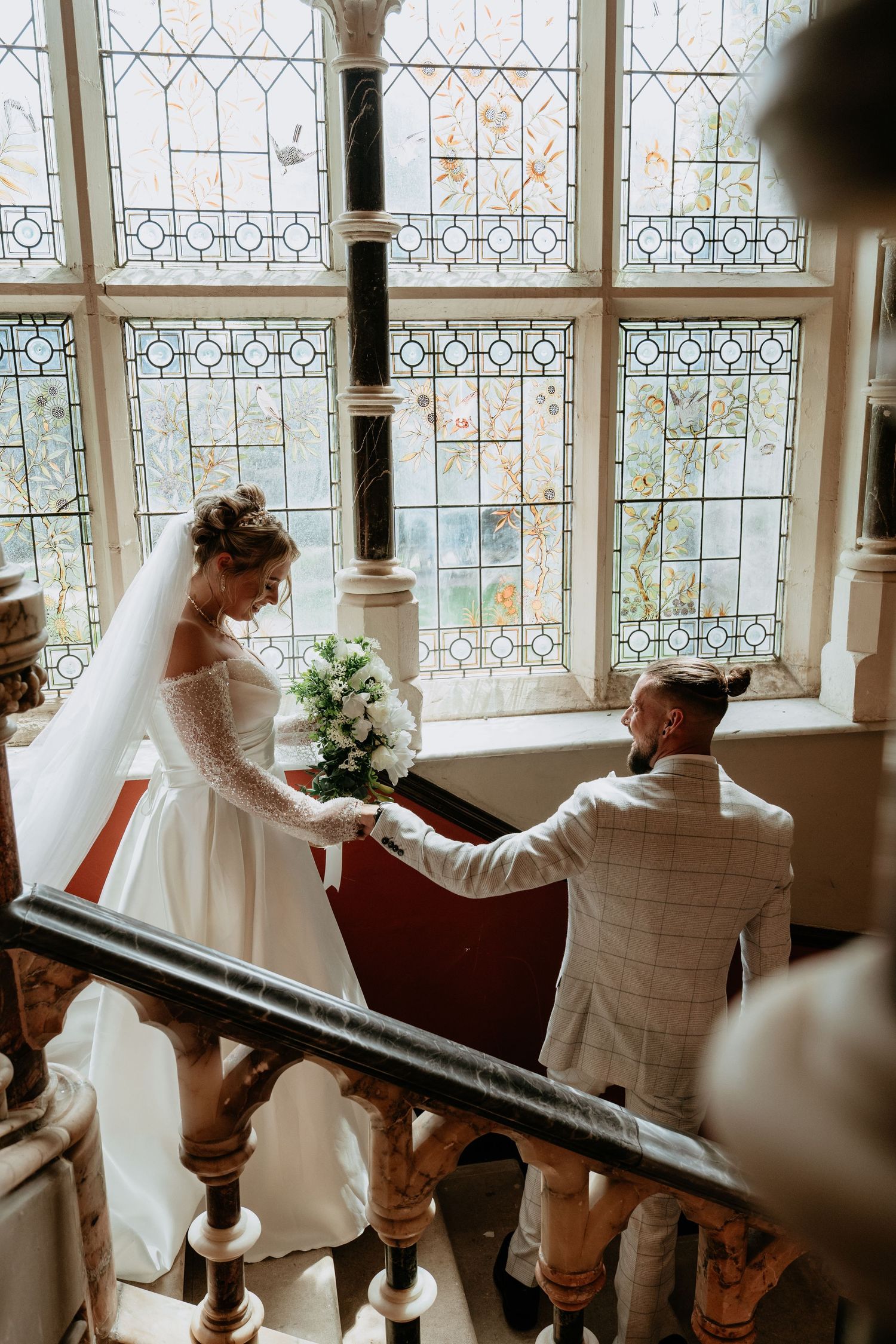Bride and groom holding hands on elegant staircase before stained glass windows in a historic wedding venue.