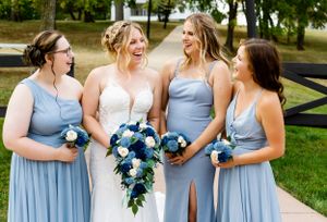 Bridesmaids in light blue dresses holding blue and white bouquets share joyful moment outdoors.