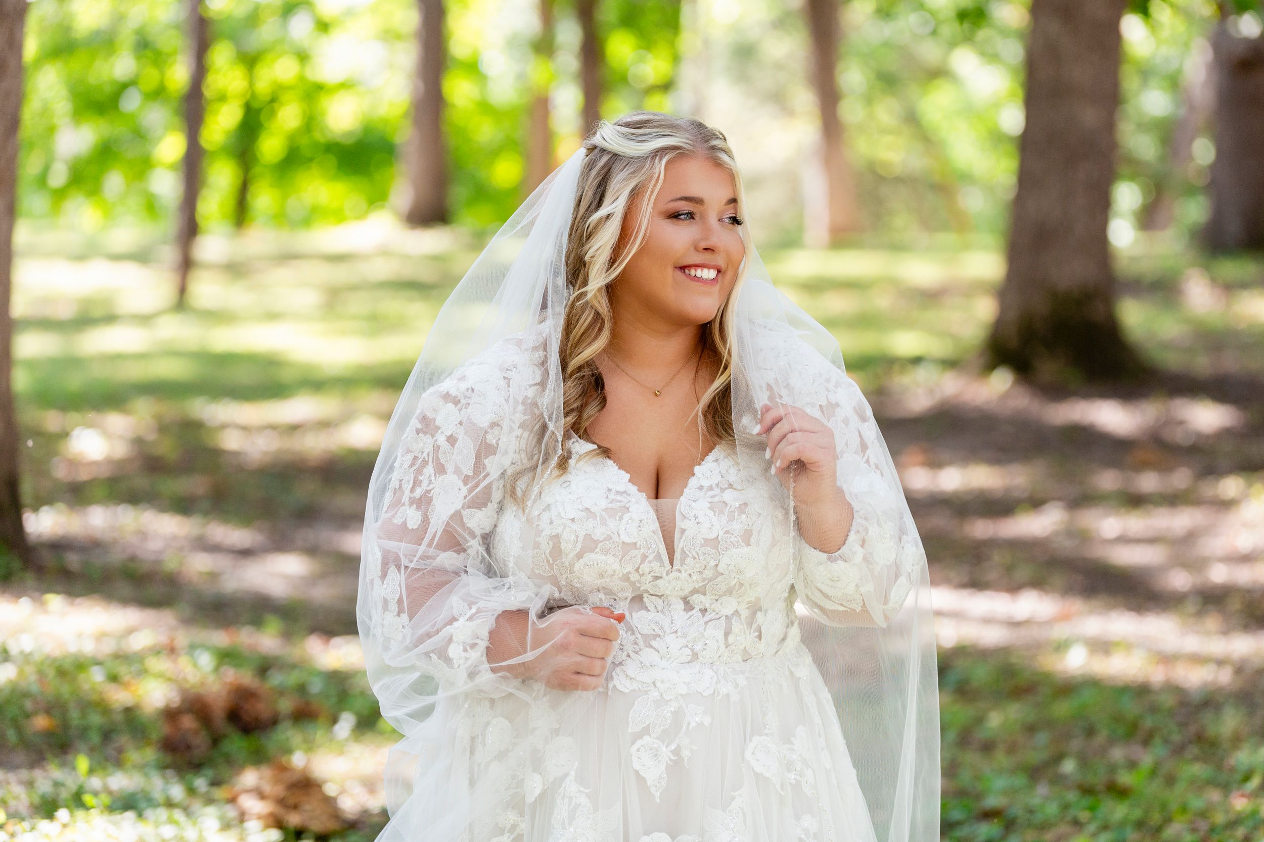 Bride in a lace wedding gown and veil smiles while standing in a sunlit forest setting.