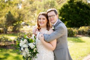 A wedding couple poses together outdoors holding a white floral bouquet.