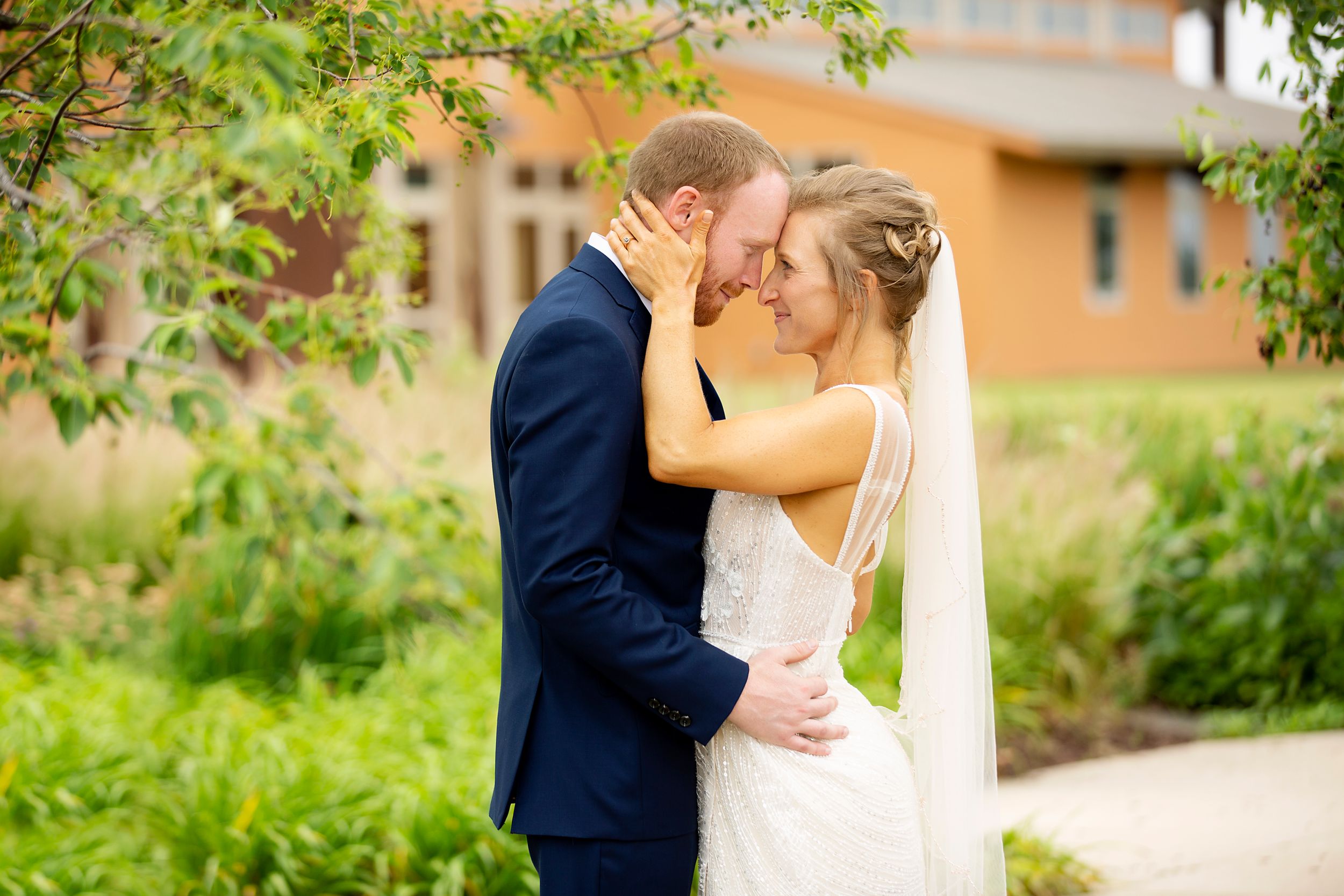 Romantic outdoor wedding kiss with greenery and warm-toned building in background.