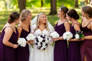 Bridal party in deep purple dresses poses together holding white bouquets outdoors.