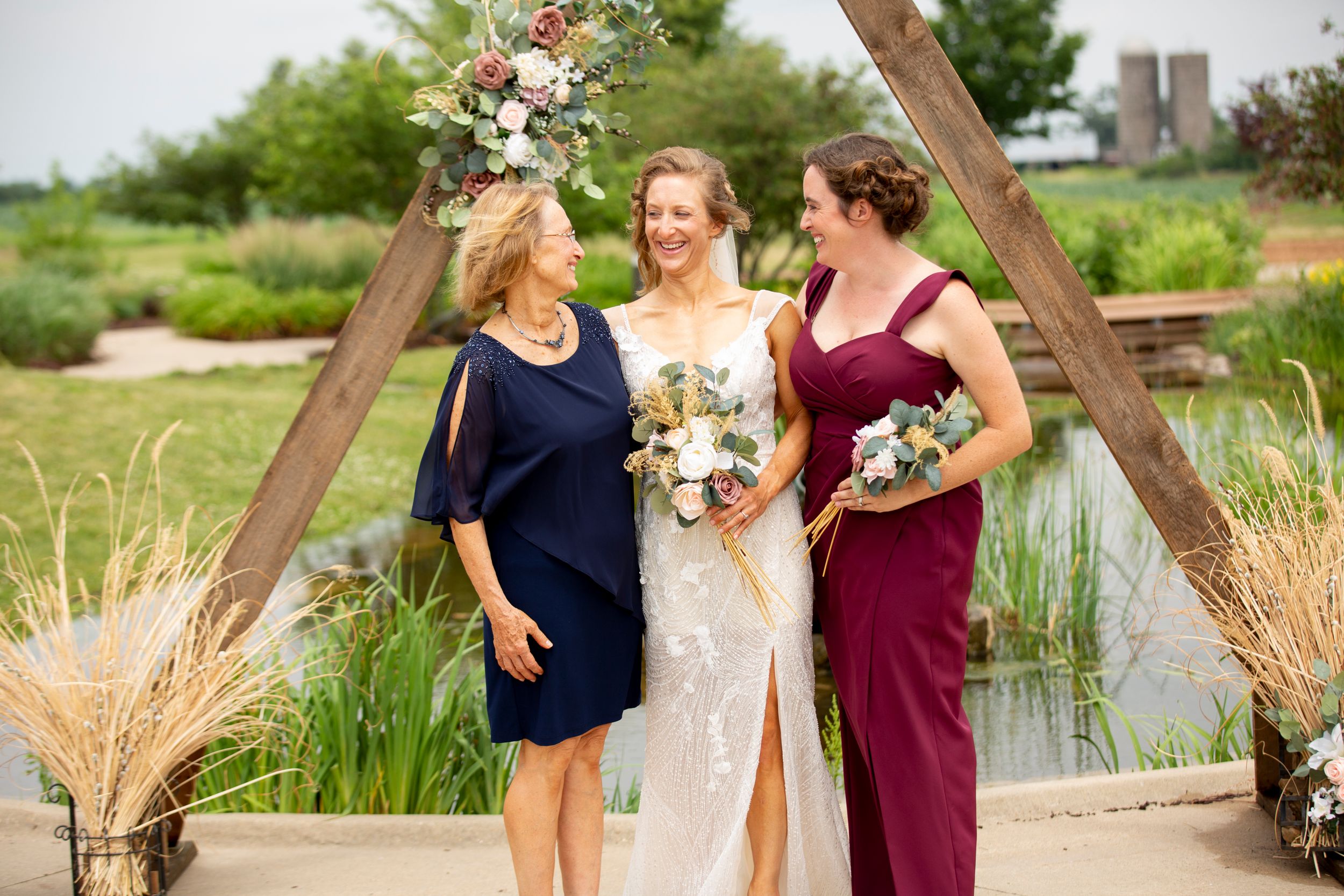 Three women in formal attire share candid laughter while posing together at an outdoor wedding venue near a pond.
