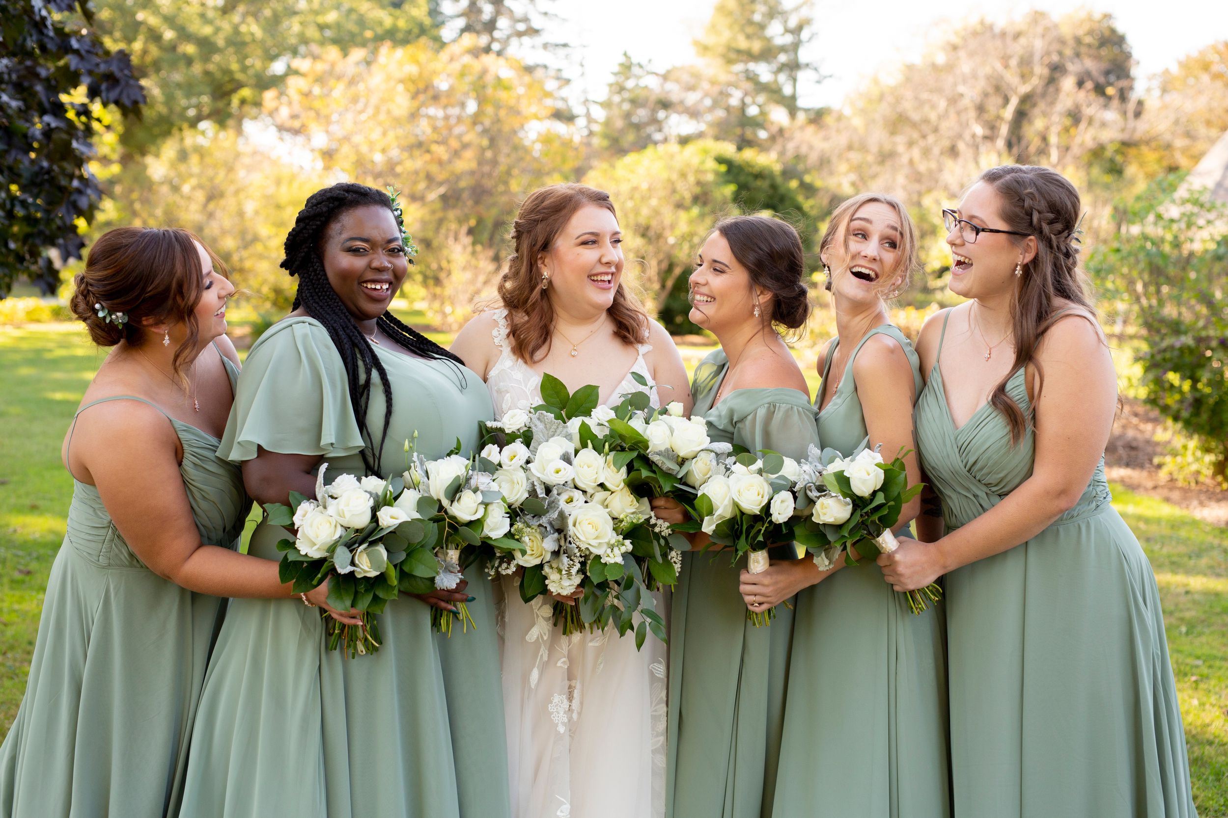 Wedding party in sage green dresses shares a candid laughing moment outdoors.
