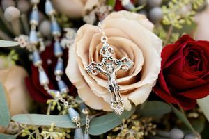 Close-up of a delicate silver crucifix nestled in a pale pink rose with red roses in the background.