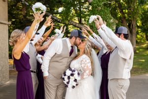 Wedding party celebrates with raised hands and bouquets under trees in a festive outdoor setting.
