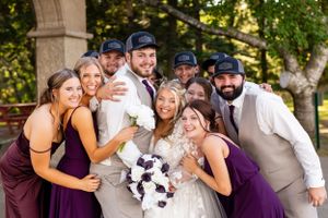 Wedding party in burgundy dresses and casual groomsmen in baseball caps pose playfully together outdoors.
