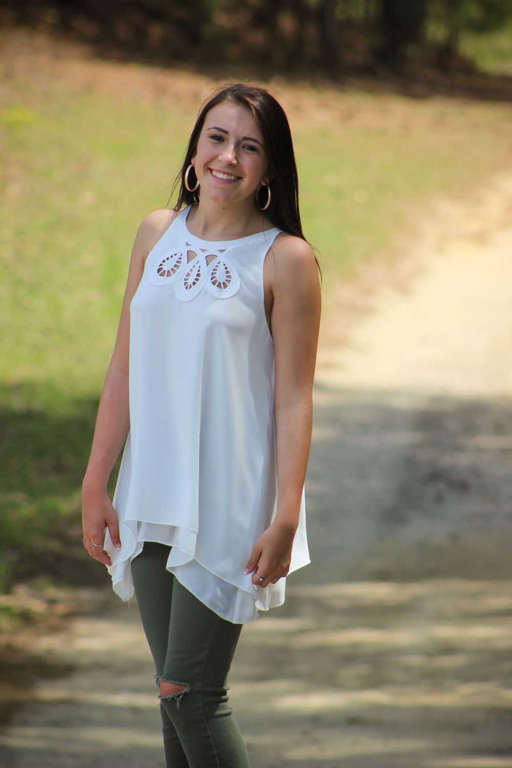 A woman wearing a flowing white sleeveless top and olive pants poses outdoors.
