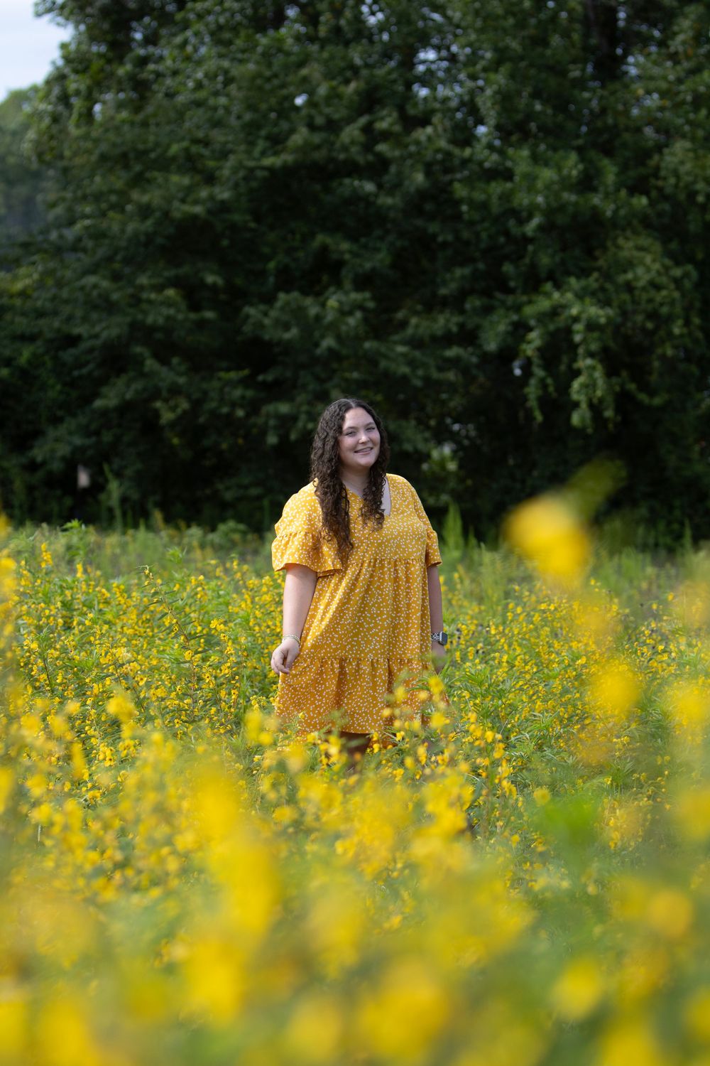 A woman in a yellow dress stands amidst a vibrant field of yellow wildflowers on a sunny summer day.
