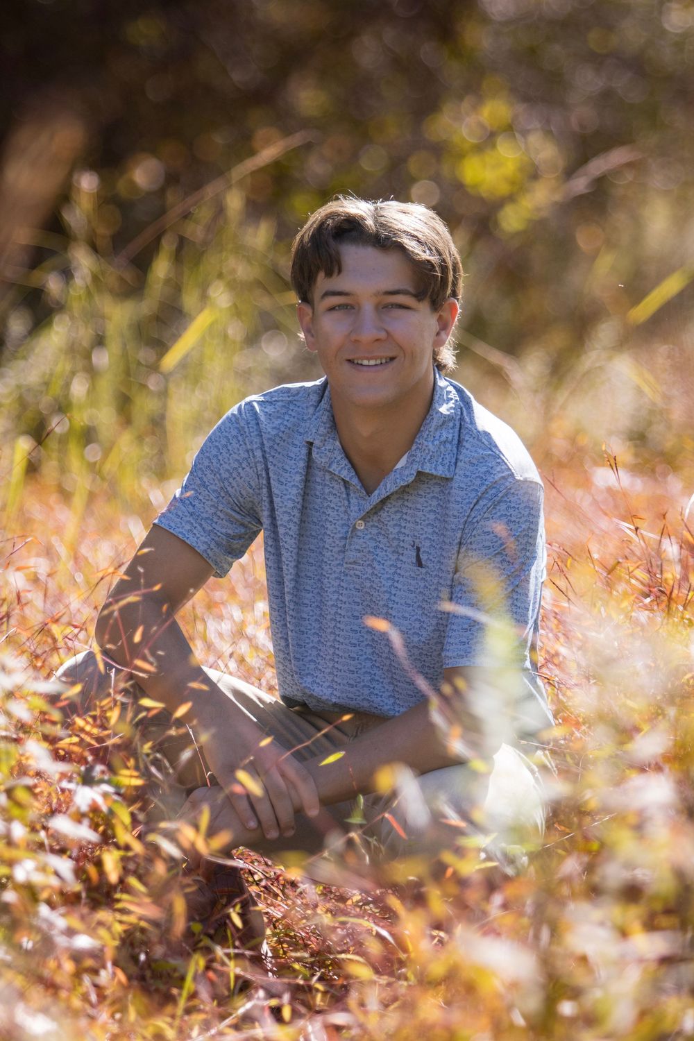 Senior in blue polo shirt lying in autumn leaves with golden sunlight filtering through trees in outdoor portrait session.