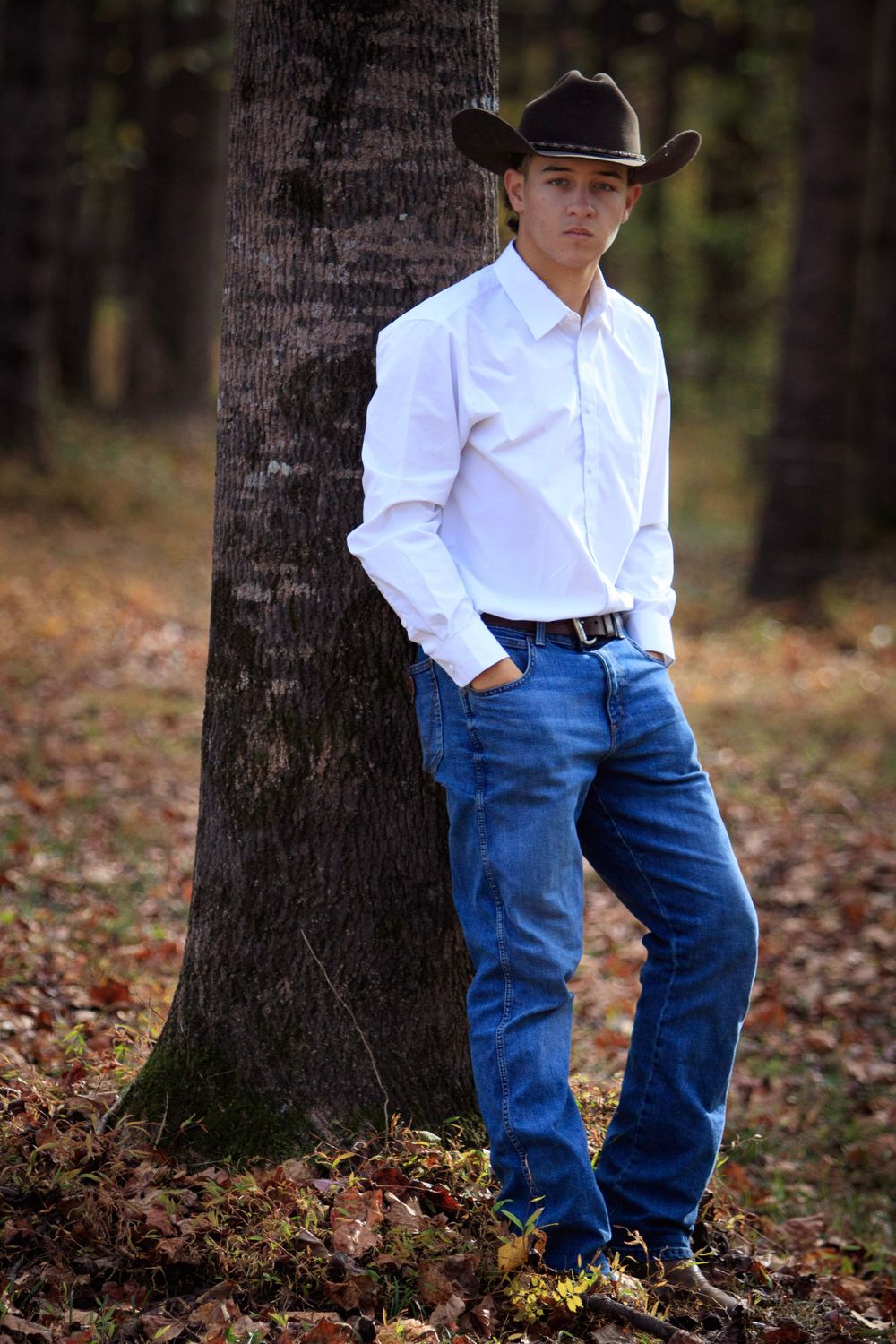 Young child in white shirt and blue jeans leans casually against a tree trunk in an autumn forest setting.