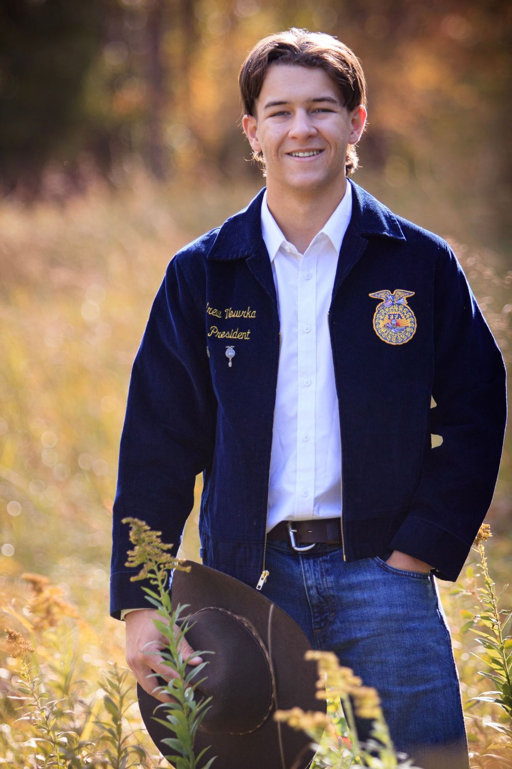 Student in FFA jacket poses in outdoor field with golden autumn foliage and natural lighting.