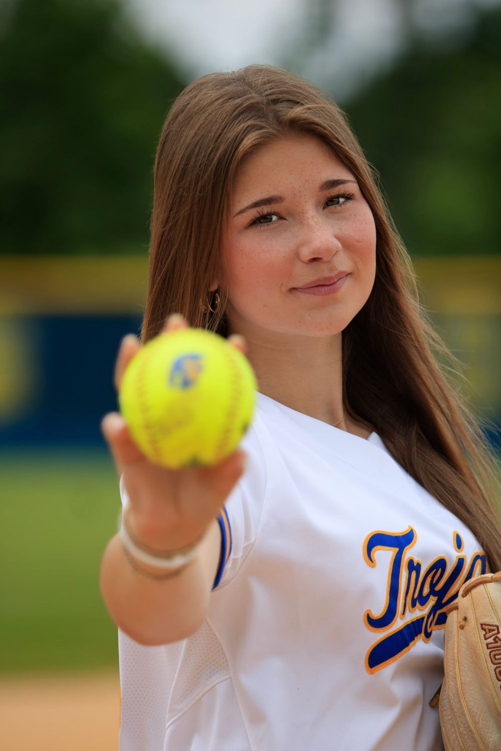 Softball player in white jersey holding up yellow softball.