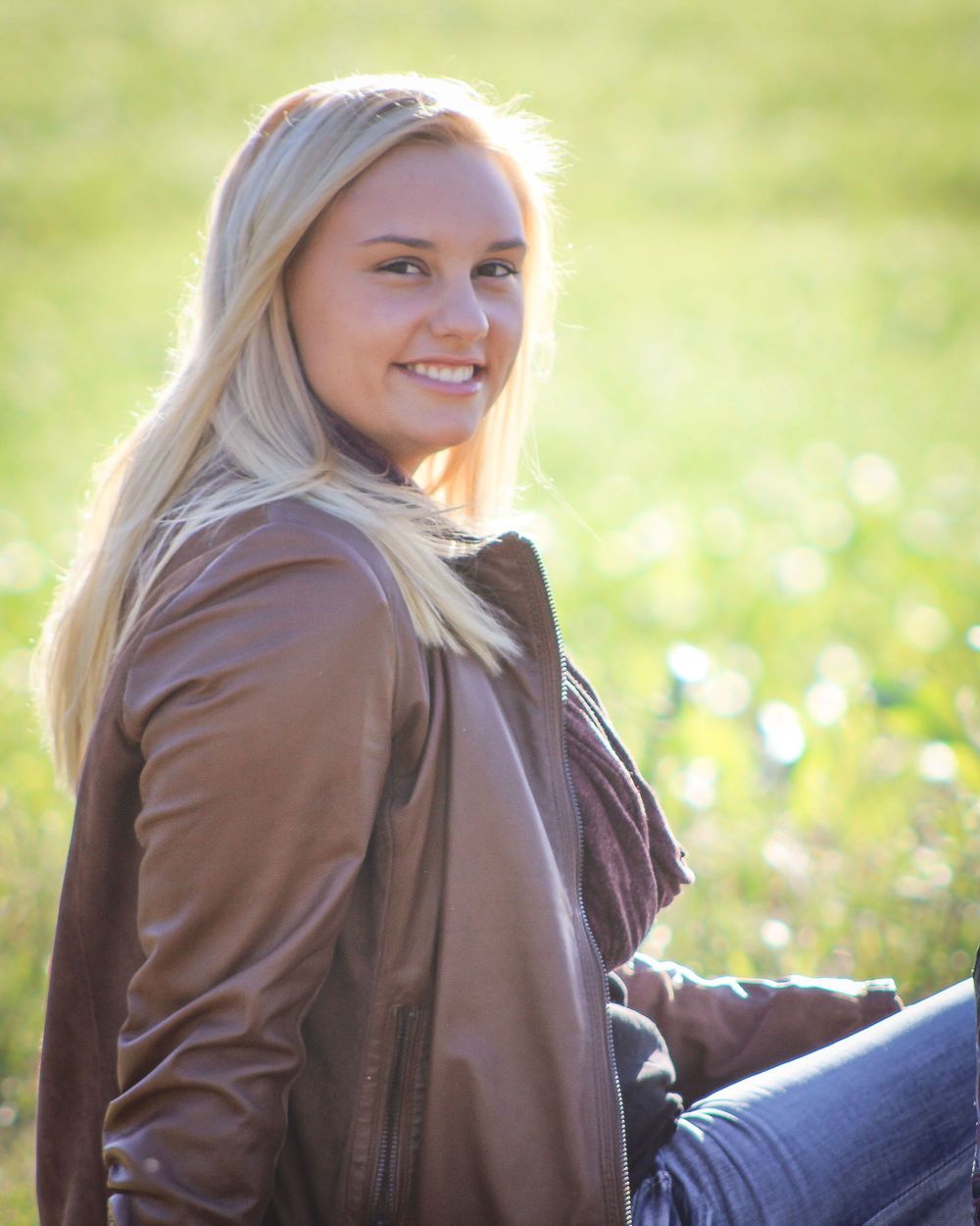 A person in brown leather jacket poses casually on grass in natural lighting.