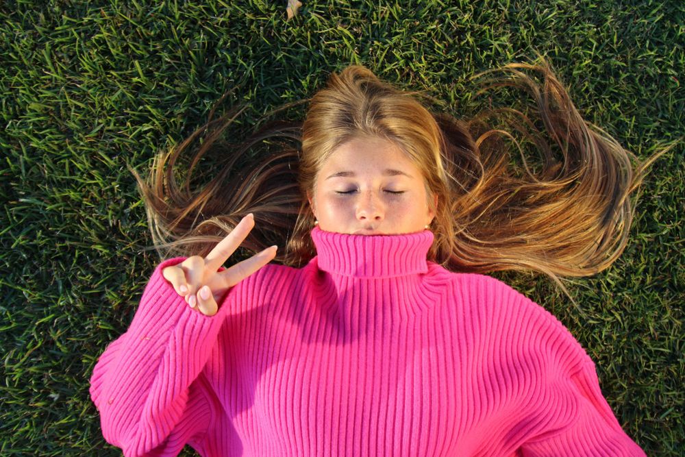 Model lying on grass wearing pink turtleneck sweater in outdoor photoshoot.