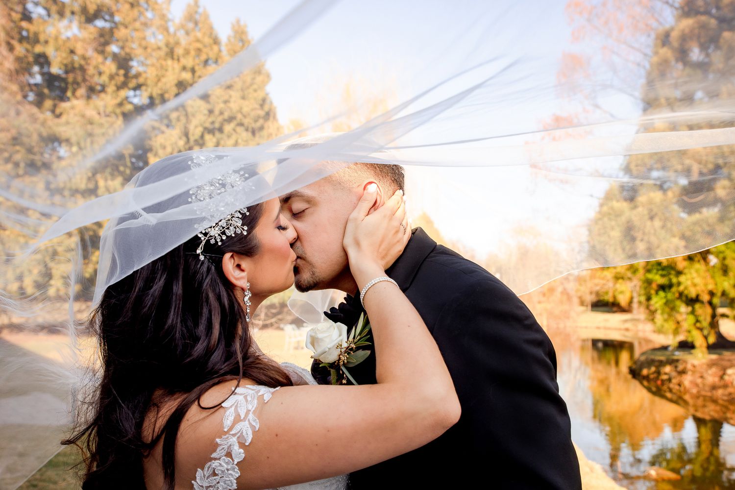 A newlywed couple shares a romantic kiss under a flowing veil with autumn trees in the background.