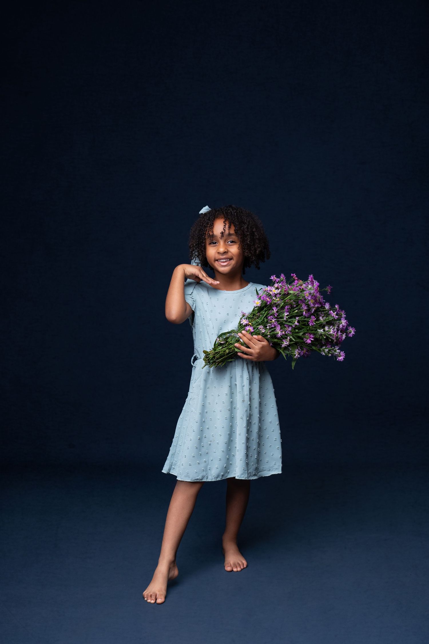 Young child in blue dress holds purple wildflowers against dark background.