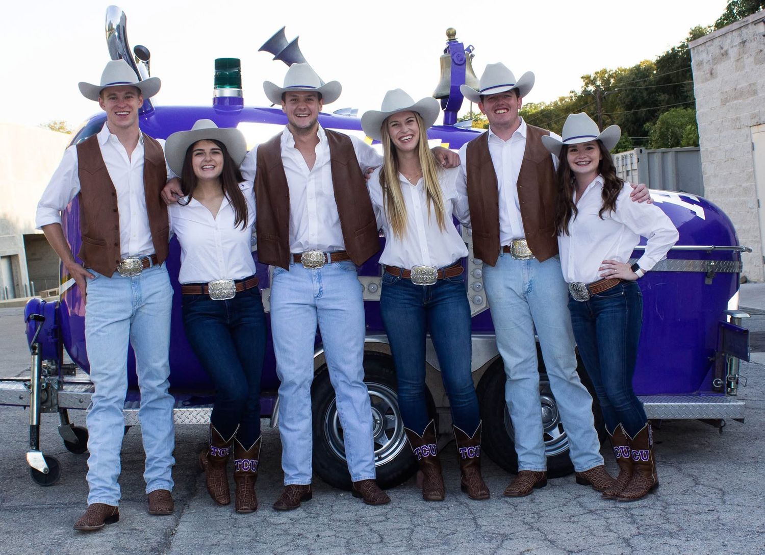 Group in western attire with cowboy hats, jeans and brown vests pose together by purple equipment outdoors.