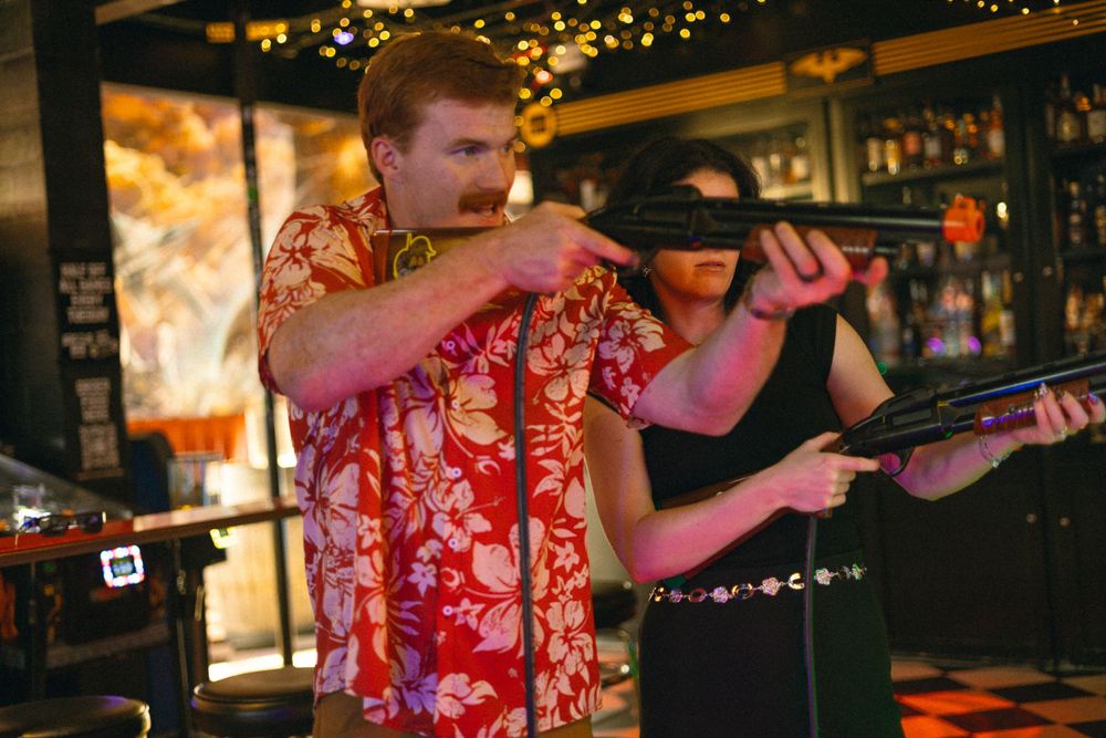 A performer in a vibrant red Hawaiian shirt plays on stage at a bar with atmospheric lighting.