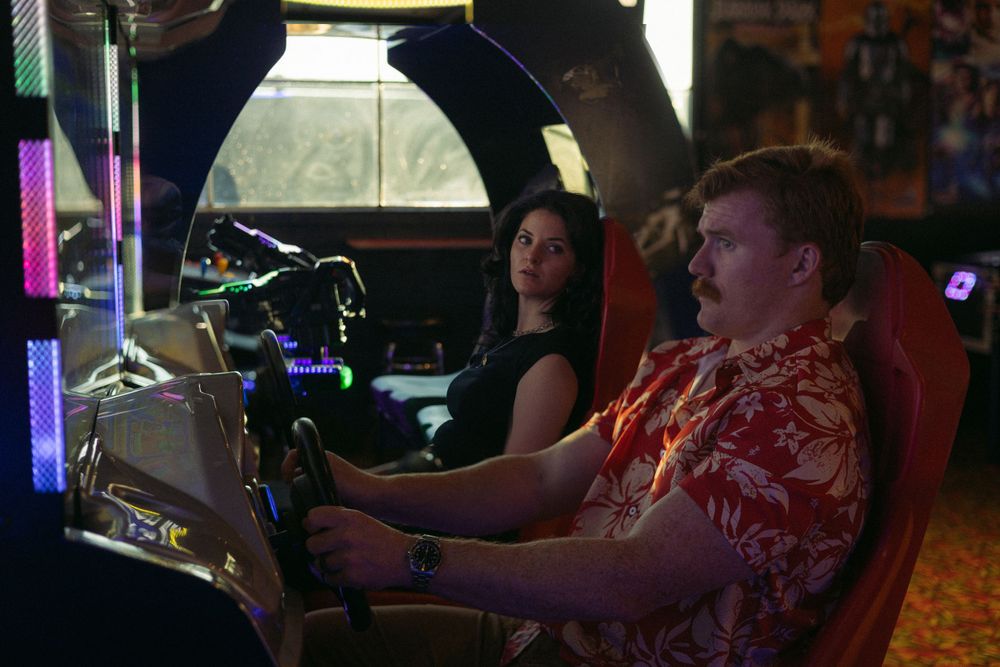 People sit at an arcade game cabinet with colorful lighting in a dimly lit entertainment venue.