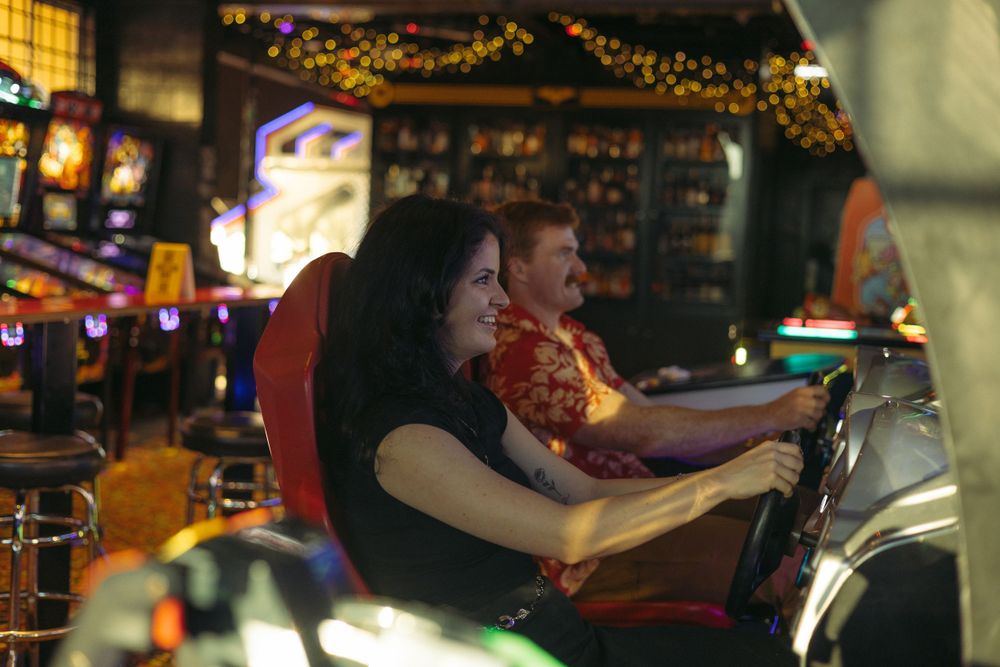 Two people sit at a dimly lit bar counter with string lights and festive decorations in the background.