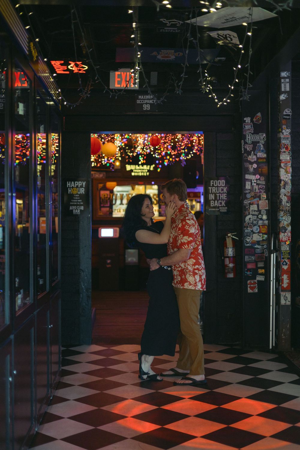Two people slow dance together in a retro diner entrance with checkerboard floors and festive lights in the background.