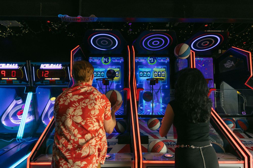 People playing arcade skeeball games illuminated by colorful neon lights at an entertainment center.