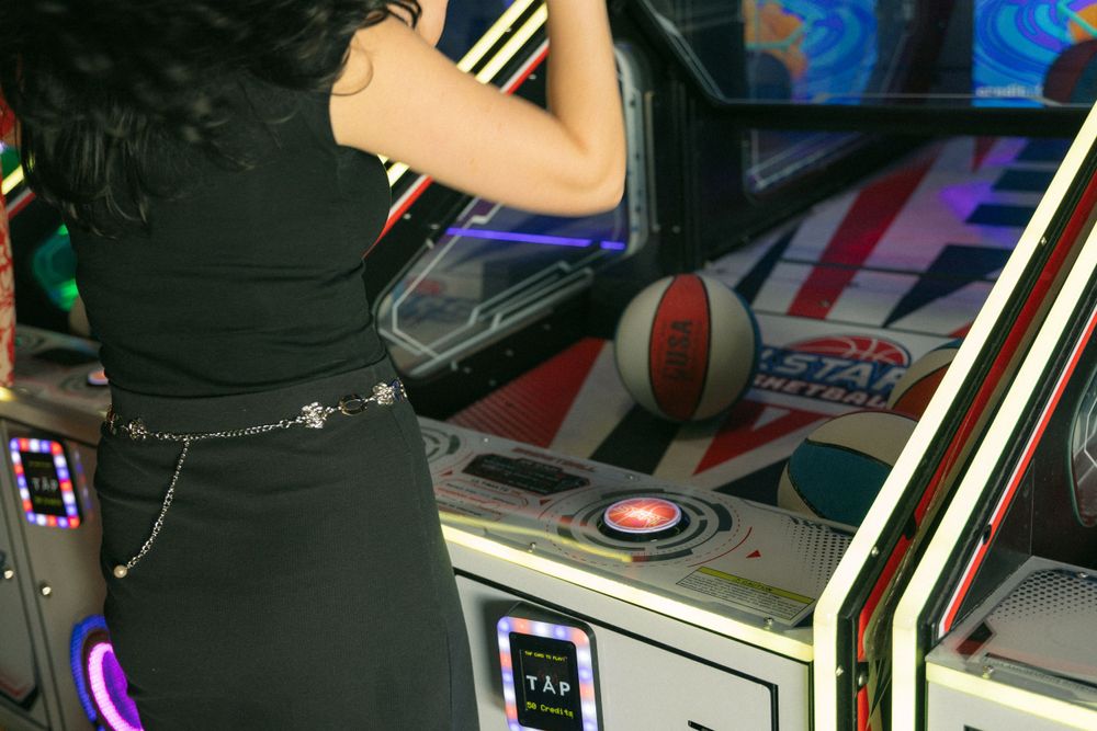 A gamer plays an arcade basketball game with neon lights and interface controls visible in the dark environment.