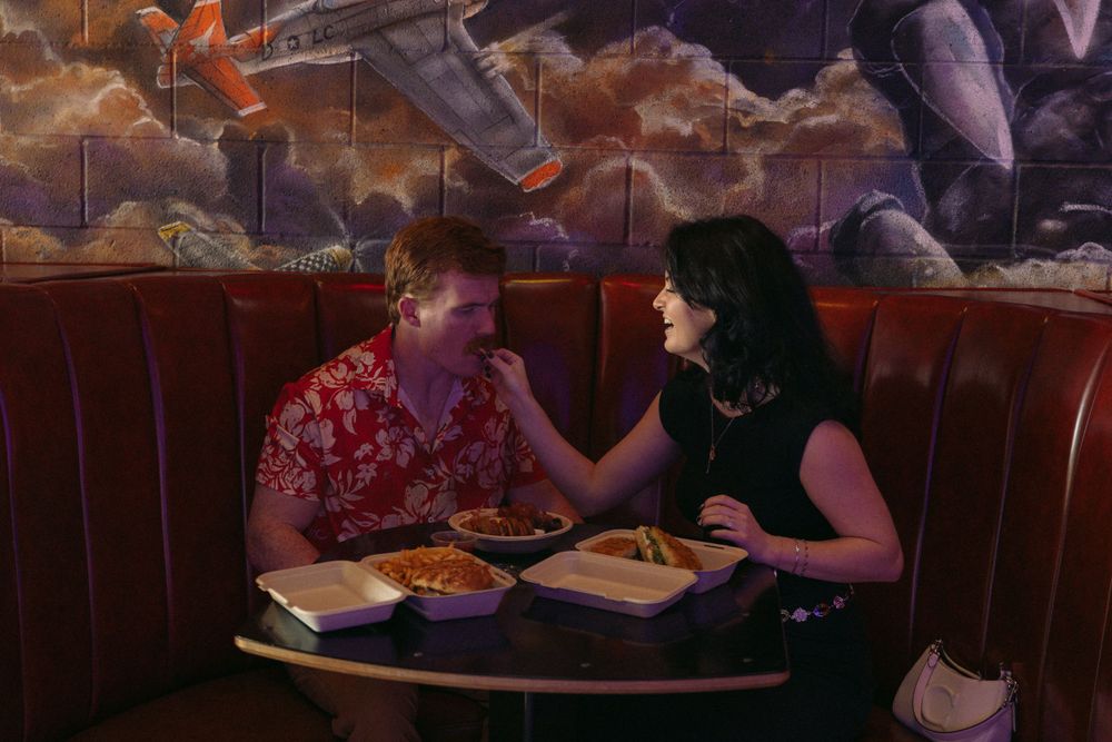 Two diners share a meal at a restaurant booth with red leather seats beneath dramatic cloudy wall murals.