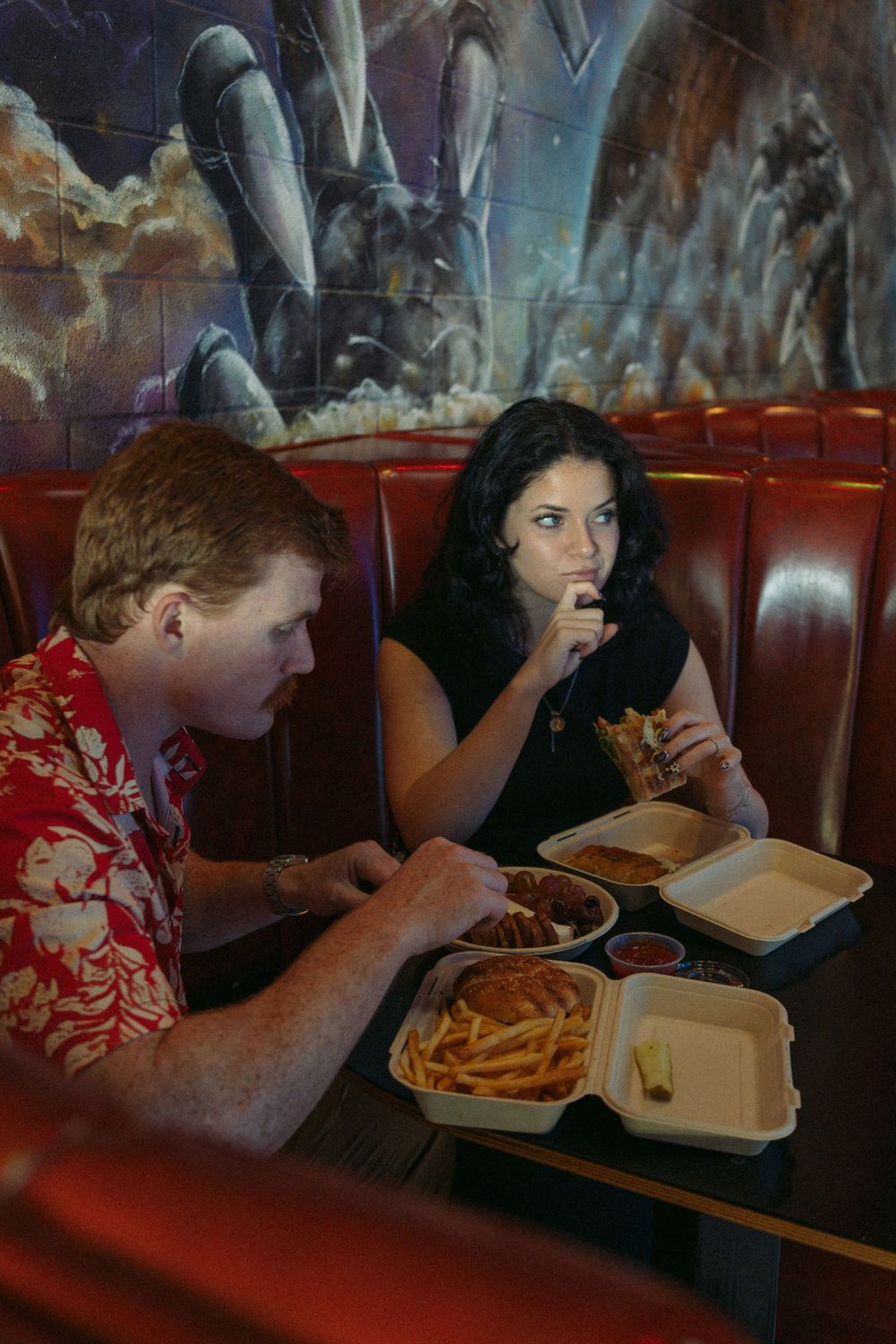 Two people enjoy a casual meal of burgers and fries in a dimly lit restaurant booth with red leather seating.