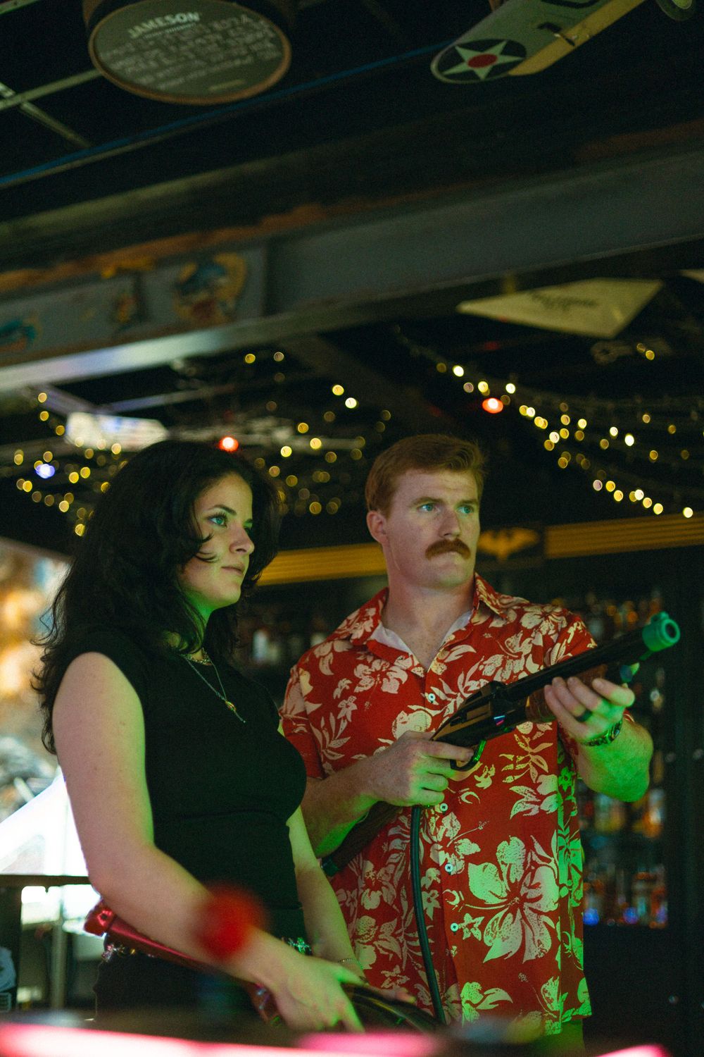 Two people stand together at a bar with string lights in the background, one wearing a bright red Hawaiian print shirt.