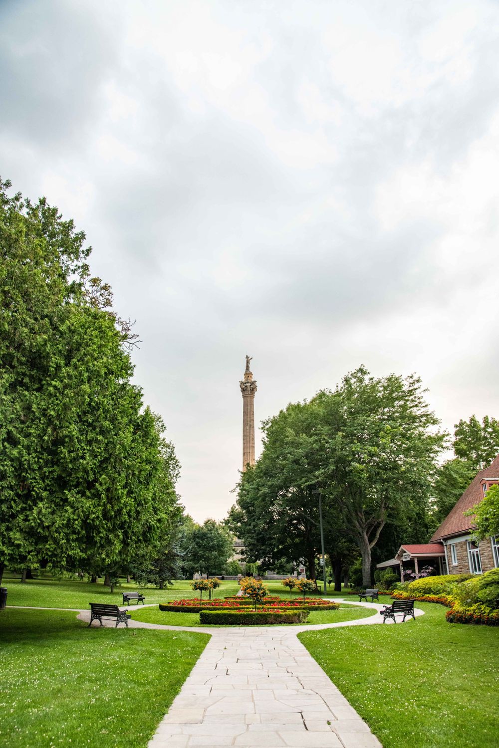 Stone pathway leading to a tall monument column in a scenic park with green lawns, trees and flower gardens.
