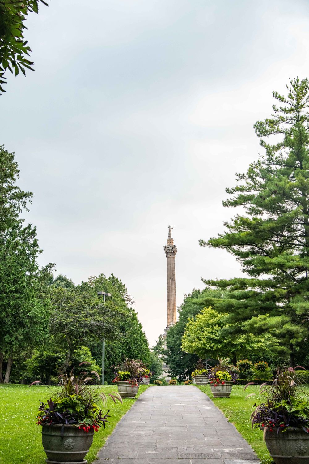 A stone pathway lined with planters leads to a tall monument column surrounded by lush green trees and landscaping.