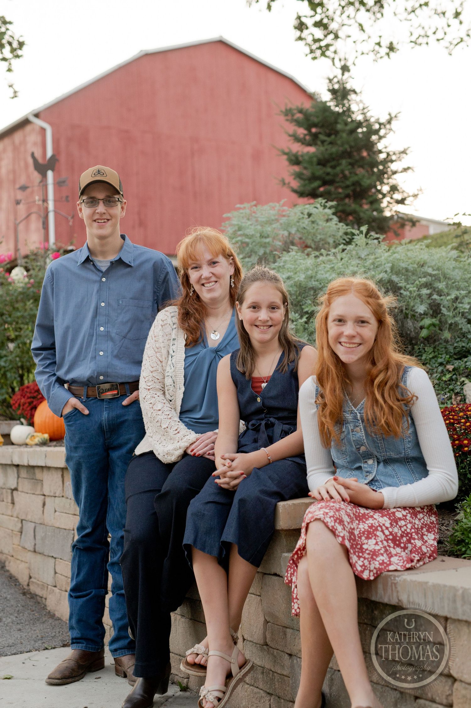 Family portrait taken on brick wall with red barn and fall decorations in background.