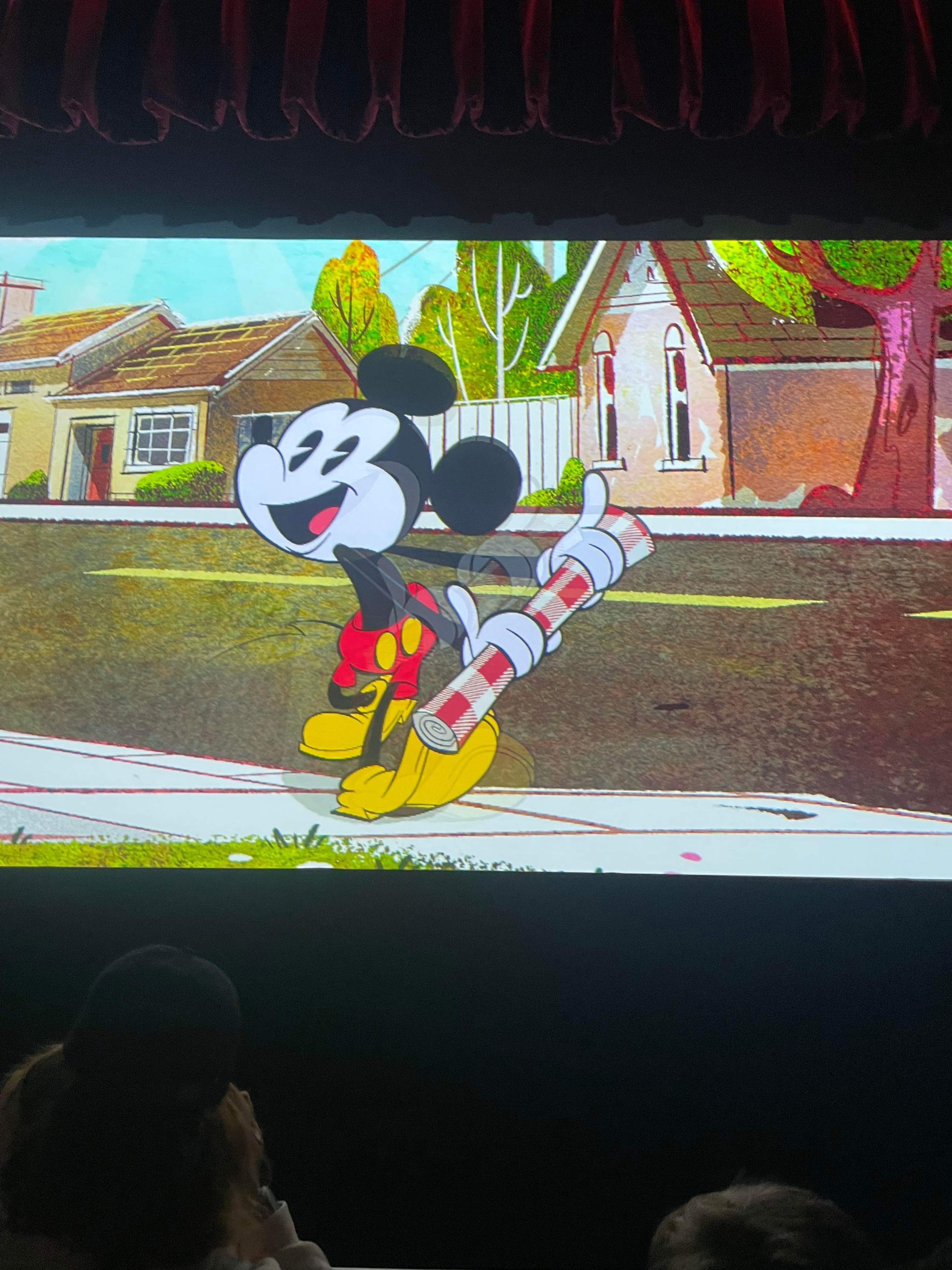 Mickey Mouse stands on a sidewalk holding a rolled up newspaper in a colorful animated neighborhood scene.