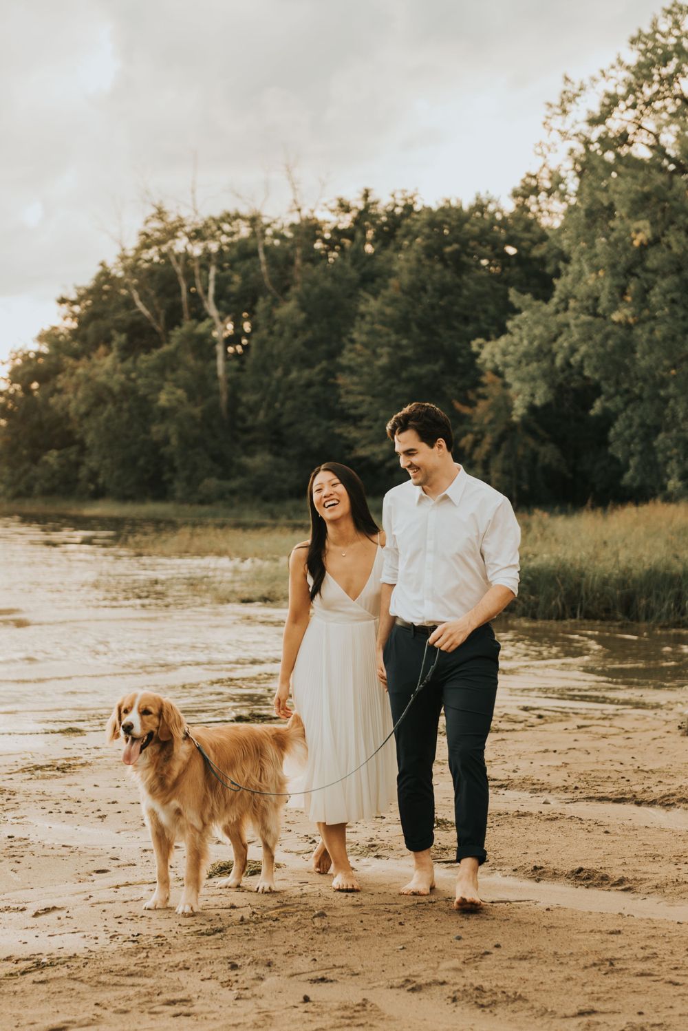 A couple walks their Golden Retriever along a sandy beach at sunset with trees in the background.