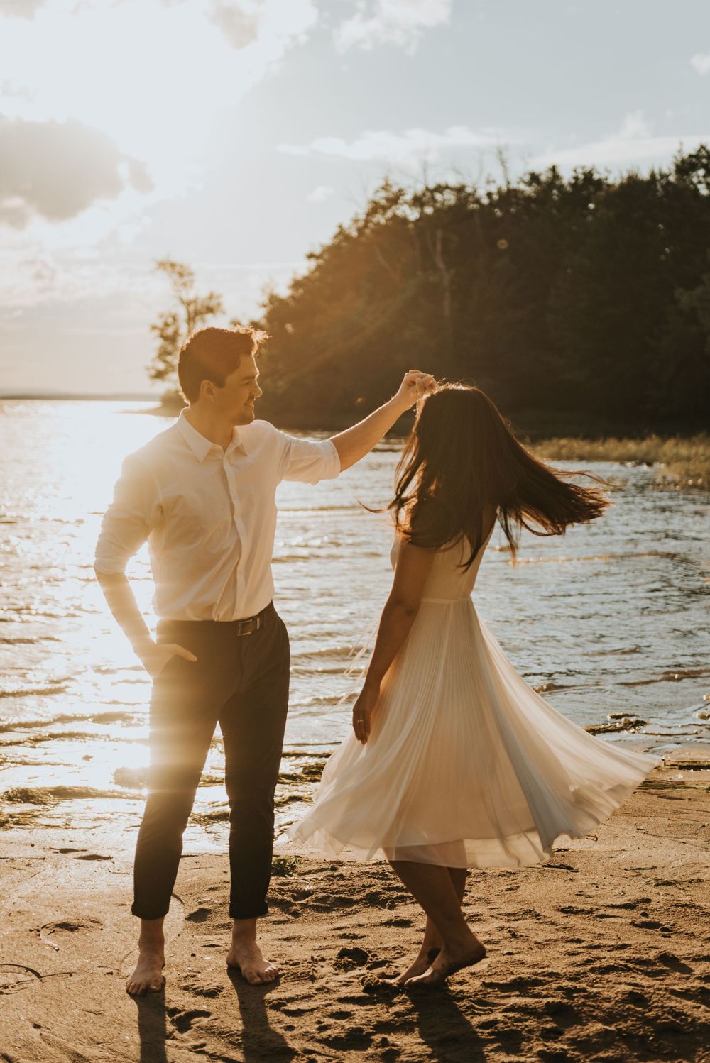 A couple dancing together on a beach at sunset with golden light streaming through trees in the background.