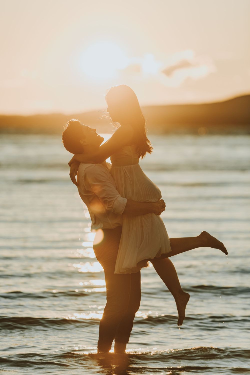 Silhouetted couple embracing in shallow ocean water during golden sunset with warm glowing sky.