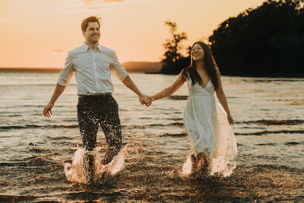 A couple splashes through shallow water at sunset on a beach, holding hands and laughing together.